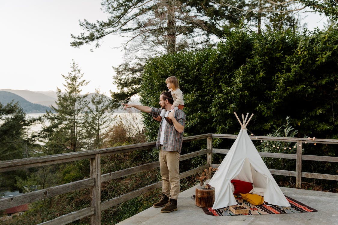 Free Side view of little child sitting on shoulders of father while man pointing to mountain while standing on wooden terrace with wigwam and picturesque view Stock Photo